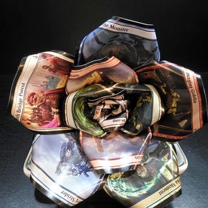 May include: A unique, handmade paper flower constructed from colorful trading cards. The flower features various fantasy art and text, including "the Monster" and "Swamp". The petals display a variety of colors, including blue, green, and brown.