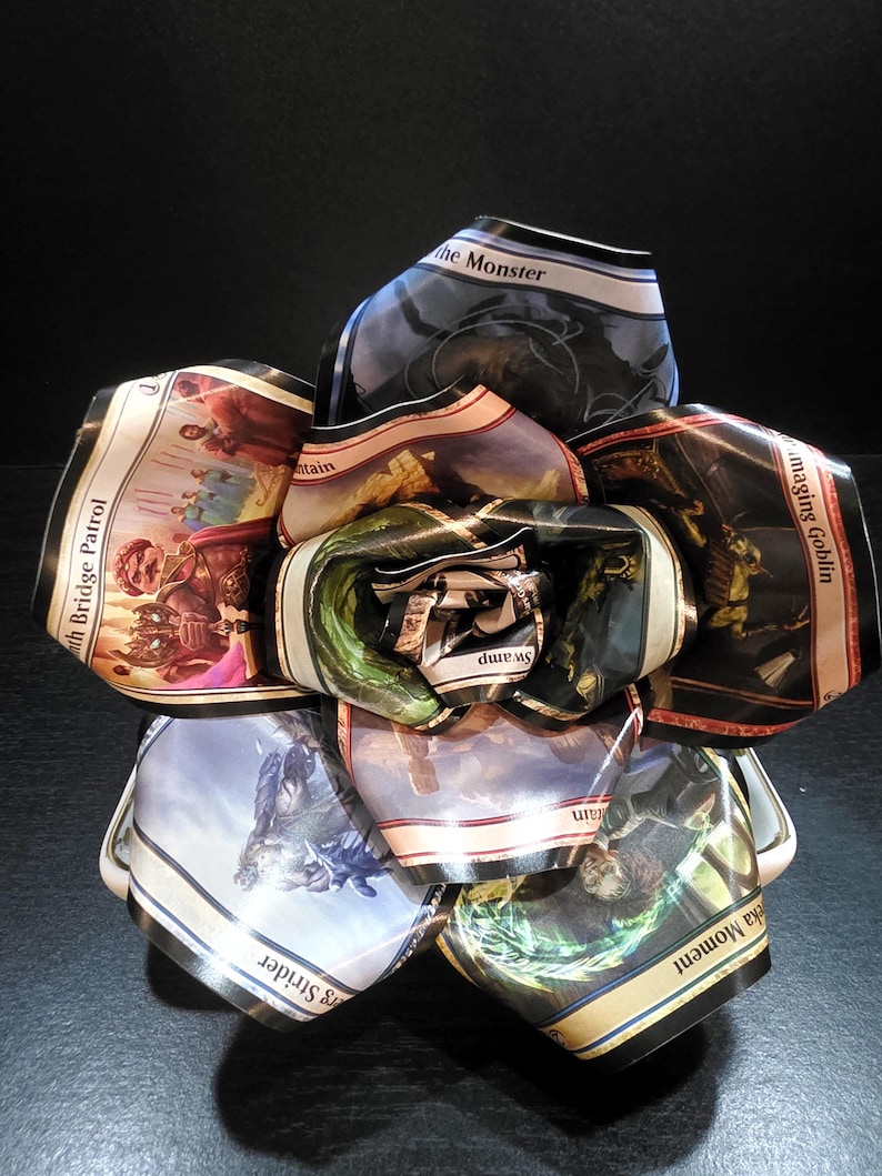 May include: A unique, handmade paper flower constructed from colorful trading cards. The flower features various fantasy art and text, including "the Monster" and "Swamp". The petals display a variety of colors, including blue, green, and brown.