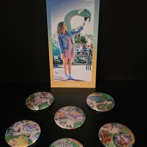 May include: A Disney's Hollywood Studios guidemap featuring a person holding a Mickey-shaped treat near a dinosaur statue, accompanied by five circular pin-back buttons displaying various illustrated park map sections and labeled locations.