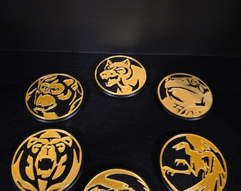 May include: A set of five circular black coasters, each featuring a raised gold-colored animal emblem. The designs include a gorilla, a tiger, a frog, a bear, a bird, and a raptor-like creature, all arranged in a circular formation.