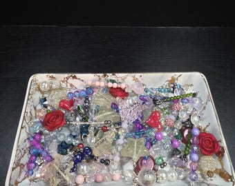 May include: A rectangular white tray with a gold marble-like pattern containing a collection of assorted beads, including red rose-shaped beads, glittery star-shaped charms, translucent spheres, and various colorful plastic and glass components.