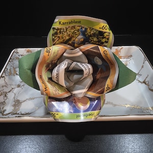 May include: A handmade paper flower made from Pokémon trading cards, featuring a variety of colors and designs. The flower is placed on a white and gold marble-patterned tray, with a dark background.