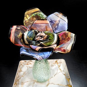 May include: A unique floral arrangement made from collectible trading cards, featuring vibrant colors and fantasy art. The cards are arranged to resemble a flower bouquet, placed in a green glass vase on a marble-patterned tray.