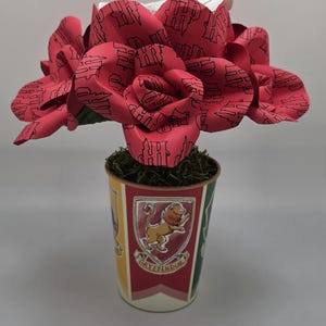 May include: A decorative paper flower arrangement featuring a large red rose with a repeating black pattern. The rose sits in a decorative cup with the Gryffindor crest. The cup is filled with green moss.