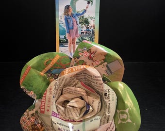 May include: A handmade paper flower crafted from Disney's Hollywood Studios park maps. The flower features green petals and a detailed center, with visible text and imagery from the maps. A park brochure stands behind the flower.