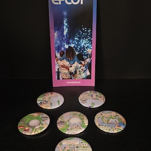 May include: A vertical EPCOT guidemap brochure with a blue and purple gradient background featuring a nighttime fireworks display and people. Below the brochure are six circular pin-back buttons, each displaying a different section of the park map.