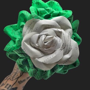 May include: A handmade bouquet featuring a large paper rose made from book pages, surrounded by green fabric leaves with a repeating black pattern. The rose has the word "CHAPTER" visible. The bouquet is held against a dark background.