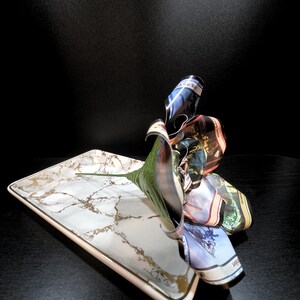 May include: A rectangular serving tray with a white marble design and gold accents. A green leaf and a cluster of folded fabric, featuring various colors and patterns, are arranged on the tray.