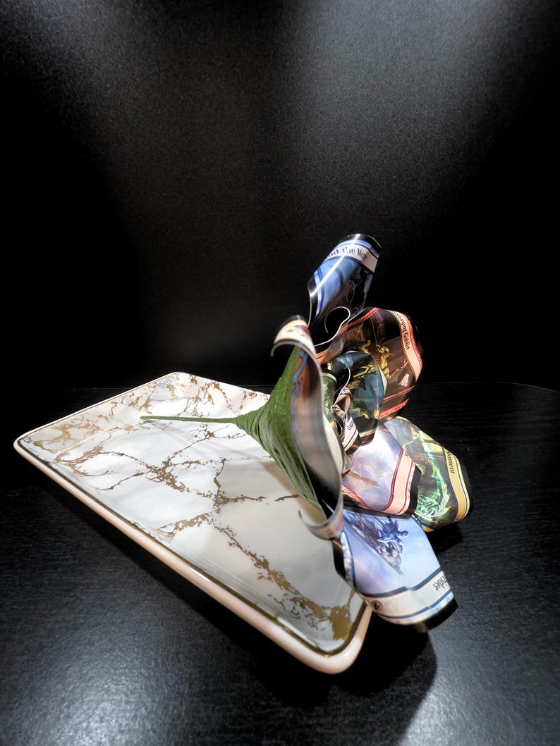 May include: A rectangular serving tray with a white marble design and gold accents. A green leaf and a cluster of folded fabric, featuring various colors and patterns, are arranged on the tray.