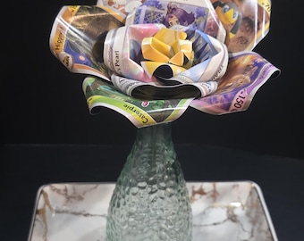 May include: A unique flower arrangement made from colorful trading cards, displayed in a textured glass vase. The vase sits on a rectangular tray with a marble design and gold trim. The flower's center is a bright yellow.