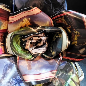 May include: A close-up of a collection of Magic: The Gathering trading card wrappers, rolled and folded to create a layered, abstract design. The wrappers display various fantasy art scenes, including mountains, swamps, and figures, with text labels.