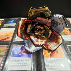 May include: A handmade flower crafted from Magic: The Gathering trading cards. The petals display various card art and text, with a mix of colors including gold, brown, and blue. The flower sits on a black background with other card-filled pages.