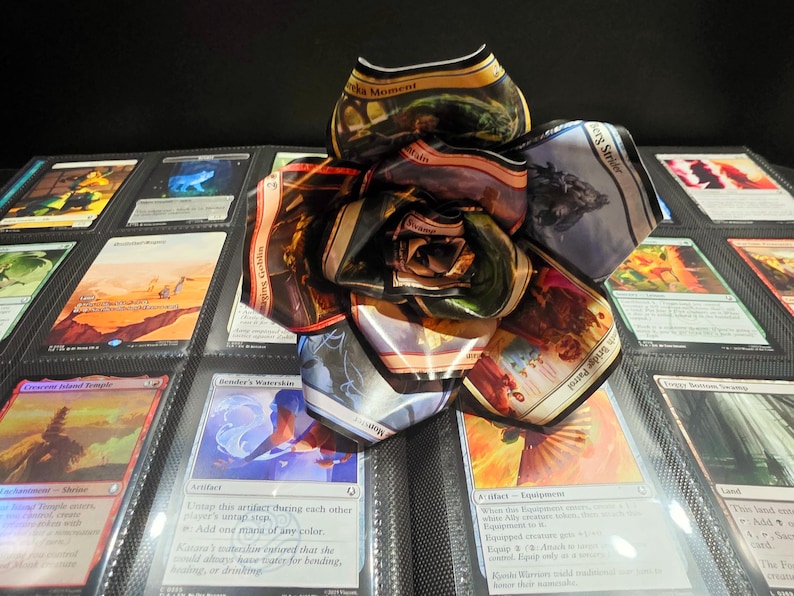 May include: A handmade flower crafted from Magic: The Gathering trading cards. The petals display various card art and text, with a mix of colors including gold, brown, and blue. The flower sits on a black background with other card-filled pages.