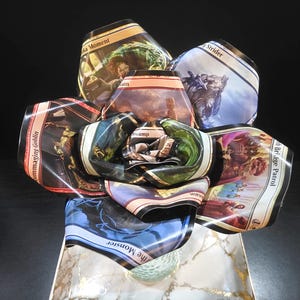May include: A unique floral arrangement crafted from Magic: The Gathering trading cards. The cards, featuring various fantasy art and text, are arranged to resemble a flower. The base is a rectangular dish with a marble and gold design.