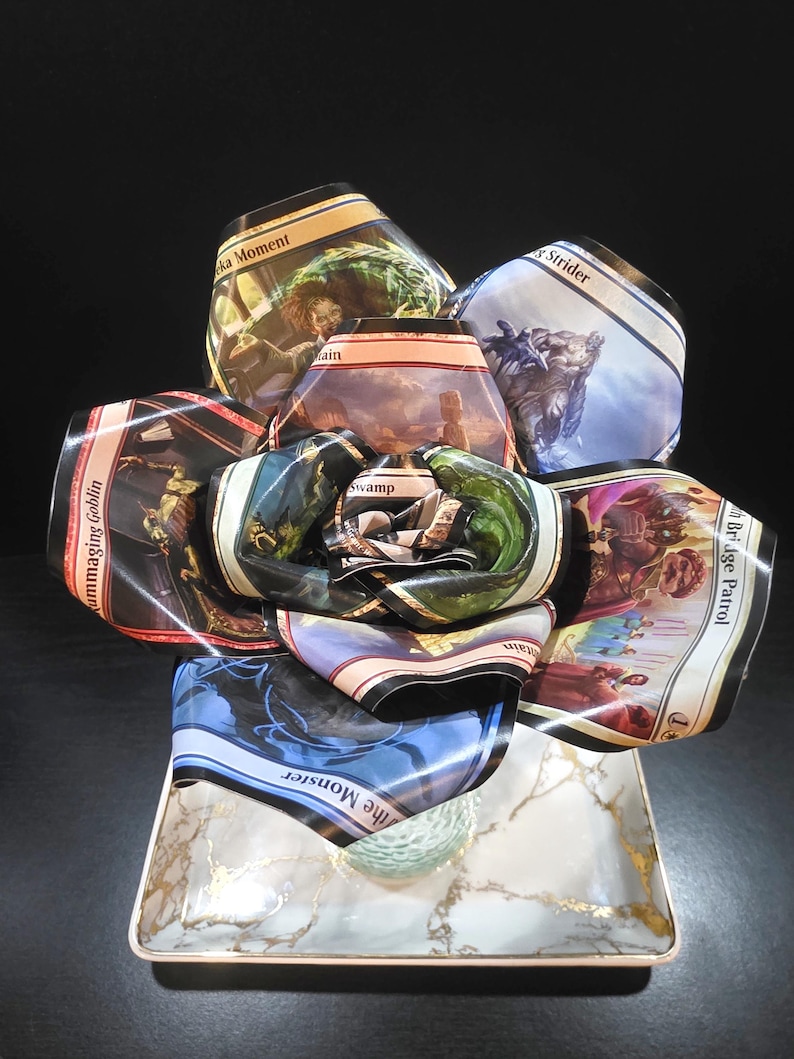 May include: A unique floral arrangement crafted from Magic: The Gathering trading cards. The cards, featuring various fantasy art and text, are arranged to resemble a flower. The base is a rectangular dish with a marble and gold design.