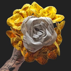 May include: A handmade paper flower bouquet. The central flower is crafted from rolled book pages, surrounded by yellow fabric petals printed with a repeating black design. The bouquet is held against a dark background.