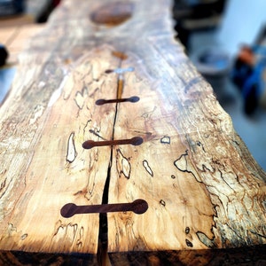 May include: A close-up of a wooden table top with a natural wood grain pattern. The table top is split in half and has four dark brown wooden dowels connecting the two halves.
