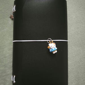 Könnte beinhalten: Ein schwarzer Einband für ein Notizbuch in Lederoptik mit einem weißen Gummibandverschluss. Ein kleiner Hello Kitty-Anhänger in einem blauen Kleid mit einem gelben Sternenstab hängt am Band. Ein silberner Sternanhänger ist an der Seite befestigt.