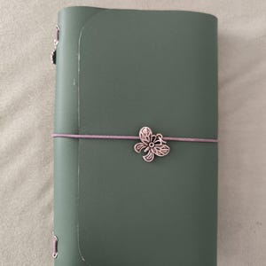 Könnte beinhalten: Ein salbeigrünes Notizbuch mit einem glatten, matten Einband. Das Notizbuch wird durch ein dünnes, hellviolettes Gummiband verschlossen, das horizontal um die Mitte verläuft. Ein silberfarbener Schmetterlingsanhänger aus Metall ist vorne am Gummiband befestigt.