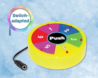 May include: A round, yellow switch-adapted device featuring a central black button labeled Push. The surface is divided into six colorful segments numbered one through six, each with a small red light. A black cable with a jack plug is attached.