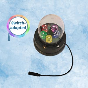 May include: A black, switch-adapted dice shaker with a clear dome. Inside the dome are six multi-colored dice. The base has a button and a black cord with a connector. The image also includes a circular graphic with the text "Switch-adapted".