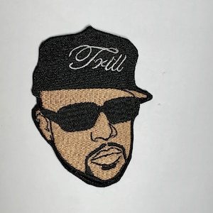 May include: Embroidered patch featuring a cartoon face wearing sunglasses and a black baseball cap with the word "T'rill" written in white on the front.