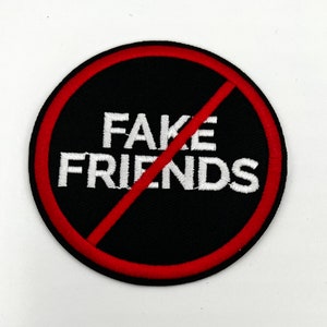 May include: Round black patch with the words "FAKE FRIENDS" in white, crossed out with a red diagonal line. The patch has a red border and is likely an iron-on or sew-on patch.