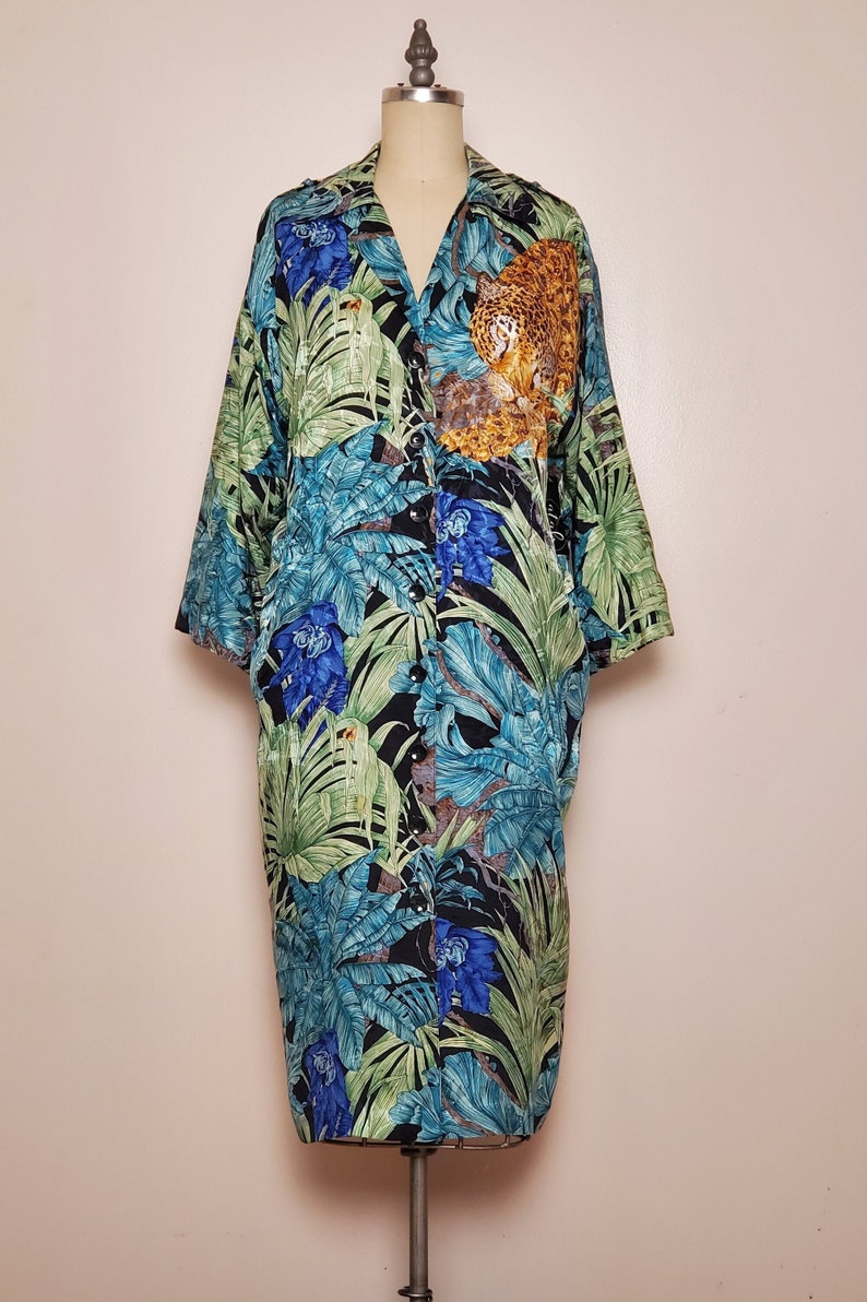 May include: A long-sleeved shirt with a tropical print featuring green, blue, and yellow leaves, a leopard, and flowers. The shirt has a button-up front and a collared neckline.