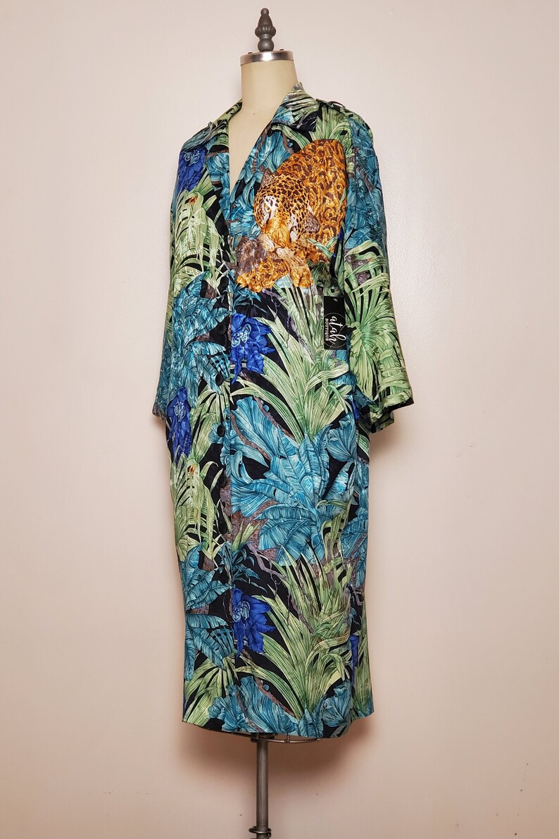 May include: A long-sleeved, button-up shirt with a tropical print featuring a leopard, parrots, and blue flowers. The shirt is on a mannequin and has a black tag that says "alah" on it.
