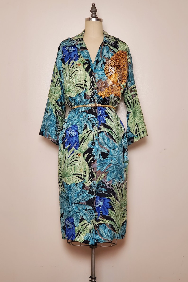 May include: A long-sleeved, button-down shirt dress with a tropical print featuring green leaves, blue flowers, and a brown and orange jaguar. The dress has a gold belt.