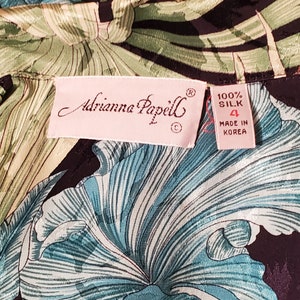 May include: A pink label with the text "Adrianna Papell" and a size 4 tag that reads "100% Silk Made in Korea". The label is on a black and green fabric with a floral pattern.