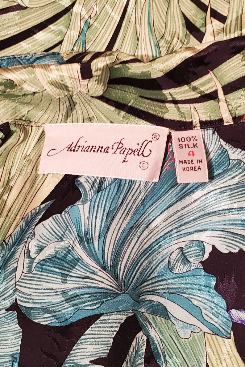 May include: A pink label with the text "Adrianna Papell" and a size 4 tag that reads "100% Silk Made in Korea". The label is on a black and green fabric with a floral pattern.