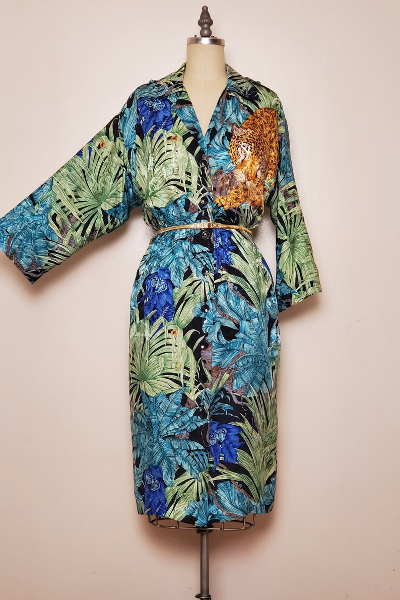 May include: A long-sleeved button-down shirt with a tropical print featuring green leaves, blue flowers, and a leopard. The shirt has a gold belt and is on a white mannequin.