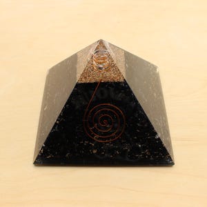 May include: A black pyramid-shaped orgone generator with a copper coil and a spiral design. The top section is a mix of gold and brown materials. The pyramid is set on a light brown surface.