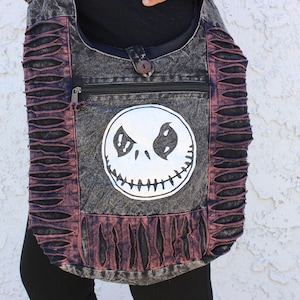 May include: A black and gray canvas shoulder bag with a distressed look. The bag has a zippered pocket and a large white Jack Skellington face graphic.