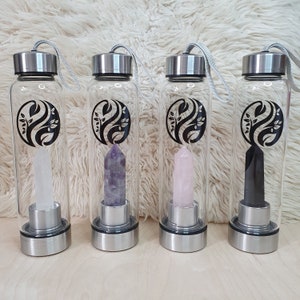 Puede incluir: Cuatro botellas de agua de vidrio transparente con tapas y bases plateadas, cada una con un cristal de diferente color: transparente, morado, rosa y negro. Cada botella tiene un diseño de hojas en blanco y negro. Las botellas están dispuestas sobre una superficie blanca y esponjosa.