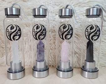 Puede incluir: Cuatro botellas de agua de vidrio transparente con tapas y bases plateadas, cada una con un cristal de diferente color: transparente, morado, rosa y negro. Cada botella tiene un diseño de hojas en blanco y negro. Las botellas están dispuestas sobre una superficie blanca y esponjosa.