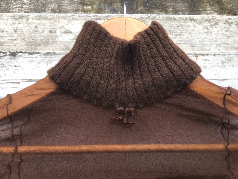 May include: A brown sheer turtleneck sweater with a ribbed knit collar. The sweater has a small embroidered logo on the front.
