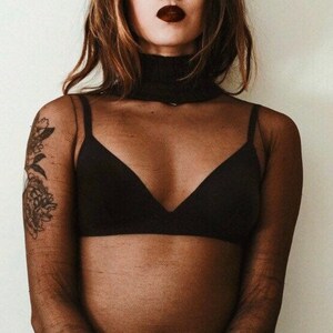 May include: A woman wearing a black sheer long-sleeved top with a black bra underneath. The top has a turtleneck style collar. The woman has a tattoo on her left arm.