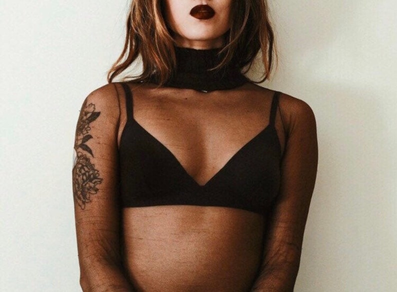 May include: A woman wearing a black sheer long-sleeved top with a black bra underneath. The top has a turtleneck style collar. The woman has a tattoo on her left arm.