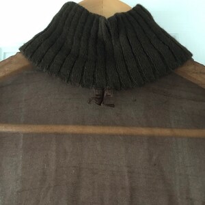 May include: A brown turtleneck sweater with a sheer overlay. The sweater has a ribbed knit texture and a small logo on the back.