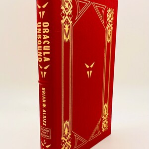 May include: A red leather-bound book with gold lettering and decorative accents. The title is "Dracula Unbound" by Brian W. Aldiss. It is a signed first edition.