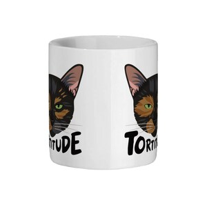 Pode incluir: Caneca de cer&acirc;mica branca com um rosto de gato preto e marrom de cada lado. O texto "Tortitude" est&aacute; do lado esquerdo e "Tortitude" est&aacute; do lado direito.