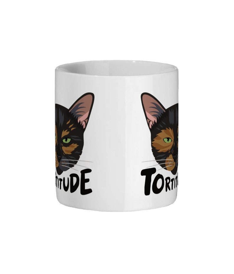 Pode incluir: Caneca de cer&acirc;mica branca com um rosto de gato preto e marrom de cada lado. O texto "Tortitude" est&aacute; do lado esquerdo e "Tortitude" est&aacute; do lado direito.