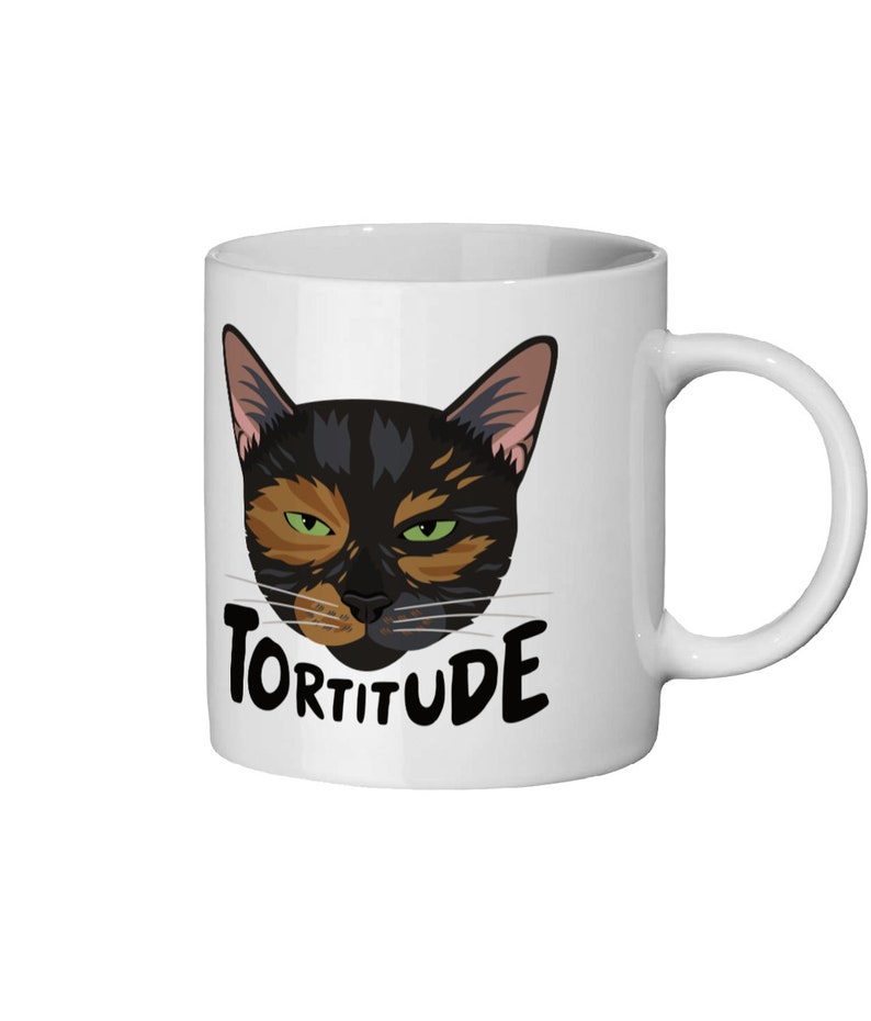 Pode incluir: Caneca de cer&acirc;mica branca com um rosto de gato preto e laranja e a palavra "Torttitude" em letras pretas.