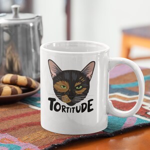 Pode incluir: Caneca de caf&eacute; de cer&acirc;mica branca com um rosto de gato preto e laranja e o texto "Tortitude" impresso nela.