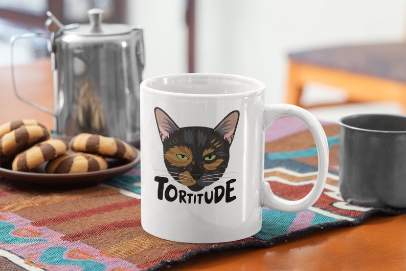 Pode incluir: Caneca de caf&eacute; de cer&acirc;mica branca com um rosto de gato preto e laranja e o texto "Tortitude" impresso nela.
