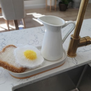 May include: A white ceramic jug with a gold tap and a white and brown knitted dish sponge shaped like a fried egg and toast.