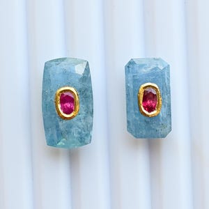 Könnte beinhalten: Zwei rechteckige Aquamarin-Edelsteine, jeweils mit einem zentralen ovalen Rubin in einer Goldfassung. Die Edelsteine werden vor einem weißen, geriffelten Hintergrund präsentiert. Die Aquamarine sind hellblau, die Rubine rot.