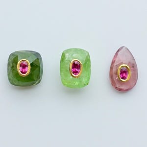 May include: Three gemstones with a central pink stone set in gold. The gemstones are in shades of green and pink, with a square, rectangular, and teardrop shape. The gems are set against a white background.